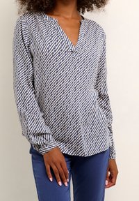 Let bluse med blå og cremefarvet geometrisk mønster, v-hals, lange ærmer og en afslappet pasform. Kombineret med blå tætsiddende bukser.