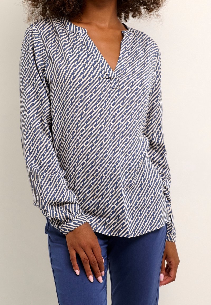 Let bluse med blå og cremefarvet geometrisk mønster, v-hals, lange ærmer og en afslappet pasform. Kombineret med blå tætsiddende bukser.