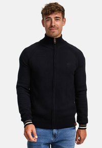 Pull noir en maille à fermeture éclair avec un col haut, des poignets et une taille côtelés, comportant un logo discret sur la poitrine et des accents rayés contrastants.