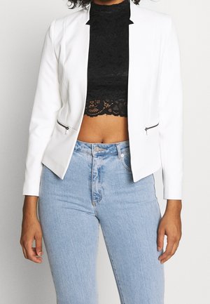 Person wearing light blue high-waist jeans, black lace crop top, and white blazer with zippered pockets, standing against white background.