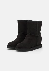 Botas de tobillo de ante negro con punta redonda, suela de cuña baja y detalle de costura decorativa. Superficie texturizada y forro suave.