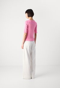 Vrouw met kort, donker haar, gekleed in een roze top met halve mouwen en wijde witte broek, staand in profiel tegen een effen witte achtergrond.