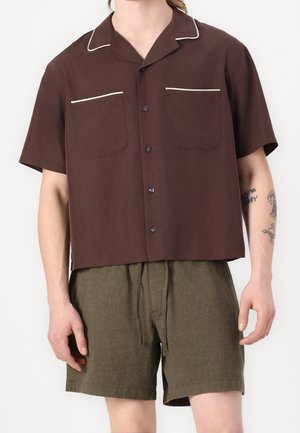 Man wearing a short-sleeve brown button-up shirt with white trim and olive green drawstring shorts, showing tattoos on right arm.