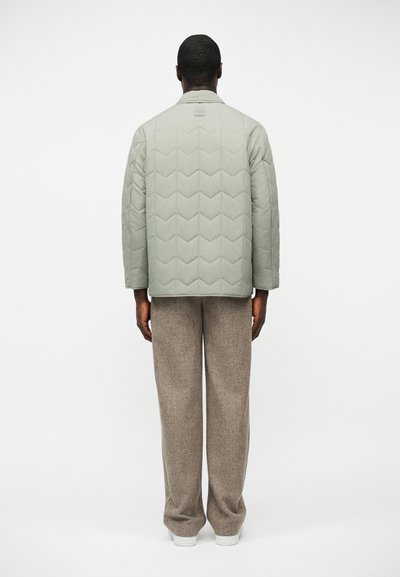 Homme de dos portant une veste matelassée gris clair et un pantalon ample beige avec des baskets blanches sur un fond uni.