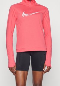 Koralfarvet langærmet top med halv-zip krave, med hvidt logo på brystet. Kombinationen består af sorte atletiske shorts, glat stof og tætsiddende design.