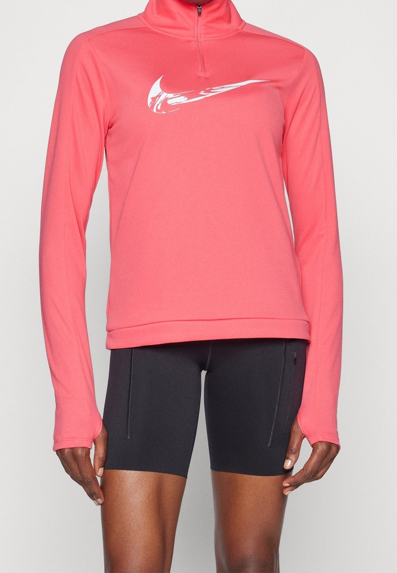 Koralfarvet langærmet top med halv-zip krave, med hvidt logo på brystet. Kombinationen består af sorte atletiske shorts, glat stof og tætsiddende design.