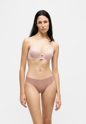 Kvinde med langt mørkt hår iført lyserød bh og mauve undertøj, står mod en ensfarvet lys baggrund.