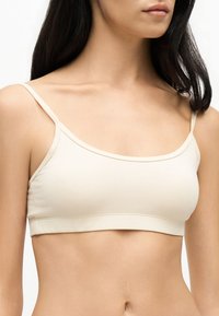 Femme portant un crop top beige clair avec de fines bretelles, montrée du cou jusqu'au haut de l'abdomen sur un fond uni.