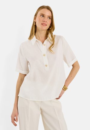 Mujer con camisa beige de manga corta y cuello con botones decorativos, pantalones beige, pendientes de aro dorados y pulseras doradas, posando de manera casual.