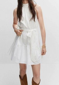 Robe blanche à col montant, taille cintrée et ceinture à nouer. Motif œillet, jupe évasée et volants superposés. Accessoirisée avec des bottes de cowboy marron.