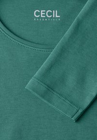 Top in misto cotone teal con scollatura rotonda, cuciture visibili e una manica. Logo del marchio "CECIL ESSENTIALS" stampato in bianco.