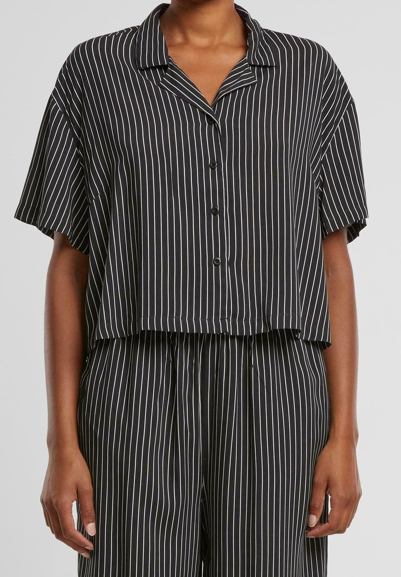 Chemise noire à manches courtes boutonnée et pantalon à fines rayures blanches verticales, portés par une personne debout, cadrés au niveau du cou et du milieu de la cuisse.