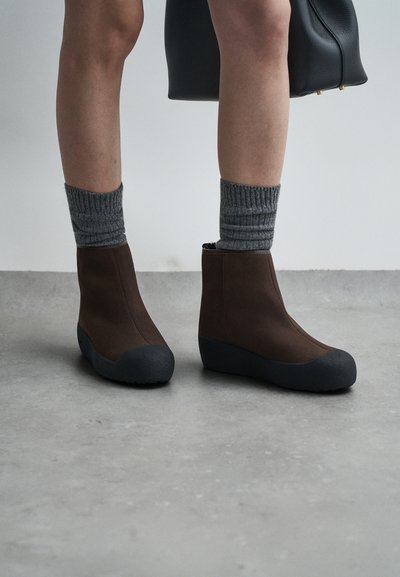 Bottes de cheville marron avec un dessus lisse, un embout en caoutchouc noir et une semelle texturée, associées à des chaussettes grises côtelées.