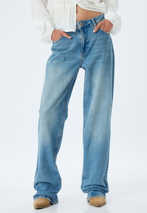 Jeans en denim bleu clair à jambe large avec une taille haute, présentant des poches avant et un lavage décoloré. Le tissu affiche une texture subtile.