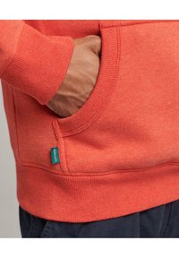 Sudadera naranja con un acabado texturizado, que cuenta con un bolsillo delantero y puños acanalados. Una pequeña etiqueta azul con "Superdry" es visible en el costado.