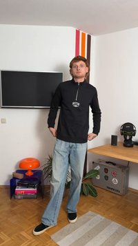 Sudadera negra con cremallera y logo de "BOSTON", combinada con jeans de pierna ancha azul claro y zapatos negros sin cordones. Entorno interior con elementos decorativos.