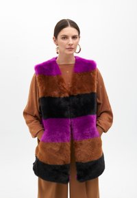 Faux fur vest with vertical stripes in purple, brown, and black. Soft texture, open-front design, paired with a brown top and gold hoop earrings.