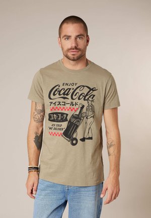 Homme portant un t-shirt beige Coca-Cola avec un graphique vintage de livraison et du texte japonais, associé à un jean bleu clair.