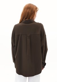 Camicia marrone a maniche lunghe con colletto, caratterizzata da una piega sul retro e una vestibilità rilassata. La consistenza appare liscia, realizzata in un tessuto leggero.