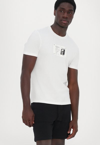 Tricou alb din bumbac, cu mâneci scurte, având o aplicație grafică pe piept. Asortat cu pantaloni scurți negri, oferind un aspect casual.