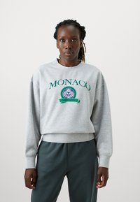 Šedá mikina s vyšívaným nápisom „MONACO“ v zelenej farbe, so zaobleným emblémom. Obsahuje rebrované manžety a lem, v kombinácii s tmavozelenými nohavicami.