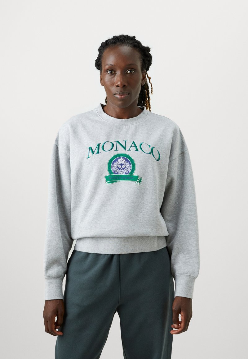 Šedá mikina s vyšívaným nápisom „MONACO“ v zelenej farbe, so zaobleným emblémom. Obsahuje rebrované manžety a lem, v kombinácii s tmavozelenými nohavicami.