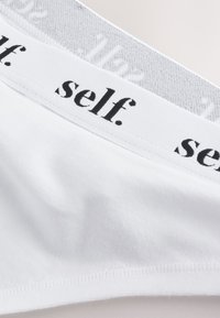 Biele bavlnené spodné prádlo s jemnou textúrou, ktoré má sivý pás s čiernym nápisom "self." Jednoduchý a minimalistický dizajn.
