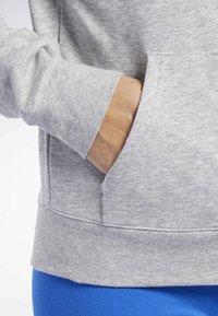 Felpa grigia con una grande tasca frontale, dettagli a coste sui polsini e sull'orlo. Tessuto strutturato con un aspetto morbido, abbinata a leggings blu.
