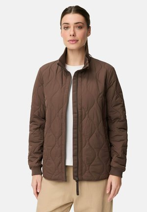 Kvinde iført en brun quiltet jakke over en hvid top og beige bukser, vendt mod kameraet med et neutralt udtryk mod en ensfarvet baggrund.