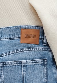 Nærbilde av baklommen på blå denimjeans med sydd brun lærlabel med teksten "DRYKORN" og beige ribbestrikket stoff over.