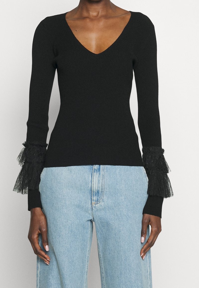 Haut noir côtelé à manches longues avec un col en V et des poignets à volants superposés en dentelle, porté avec un jean taille haute bleu clair sur une personne à la peau foncée.