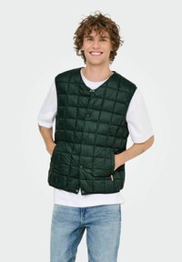 Gilet matelassé vert avec une fermeture à boutons, doté de deux poches latérales, porté sur un t-shirt blanc. Design simple et rembourré.