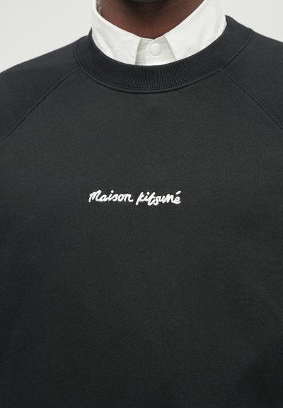 Musthave mustriga varrukatega musta dressipluusi esiküljel on valge tikandiga tekst "Maison Kitsuné". See on valmistatud pehmest kangast, millel on sile tekstuur.