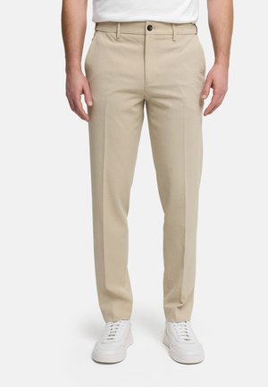 Mand iført beige skræddersyede bukser og hvide sneakers, stående mod en ensfarvet hvid baggrund.