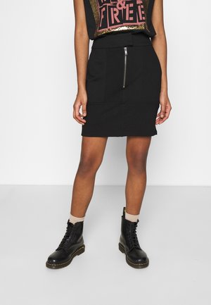 Mujer con mini falda negra con cremallera frontal, top sin mangas con diseño gráfico, calcetines beige y botas negras con cordones, de pie sobre un fondo liso.