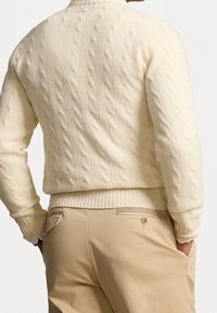 Krämsfärgad kabelstickad tröja med ribbade fållar och manschetter, visad tillsammans med beige byxor med bakficka och knäppdetalj.