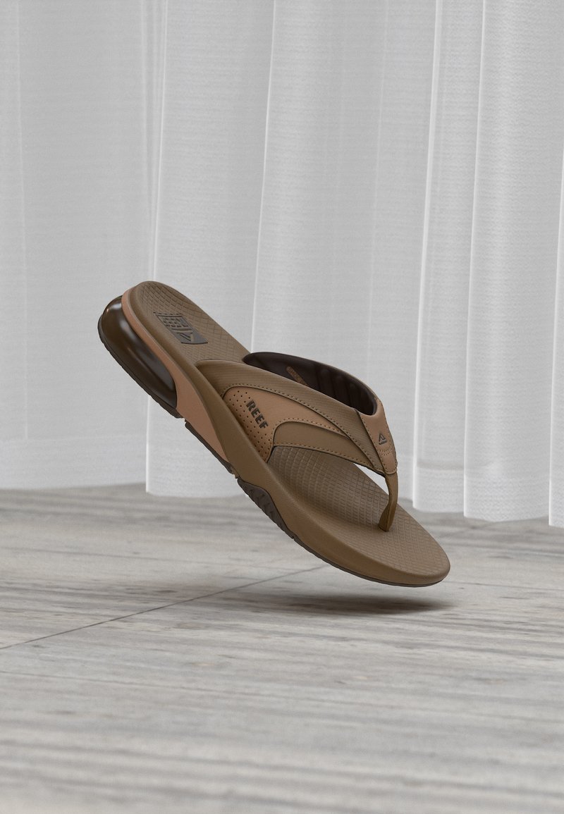 Σανδάλι flip-flop της μάρκας Brown Reef με μαξιλαρωμένο τακούνι και υφασμένο πέλμα, τοποθετημένο στον αέρα μπροστά από ανοιχτή κουρτίνα και ξύλινο πάτωμα.