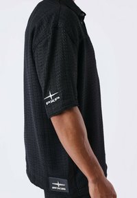Zwart short-sleeve shirt van een gestructureerde stof, met een knoopkraag en een wit logo op de linkermouw en de onderste zoom.