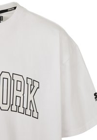 Balts kokvilnas T-krekls ar melnu "NEW YORK" teksta grafiku, īsām piedurknēm un brīvu piegriezumu. Ietver logotipa akcentu uz piedurknes.