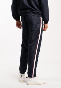 Pantaloni da jogging navy con strisce laterali bianche e rosse, vita elastica e caviglie a costine, abbinati a scarpe da ginnastica bianche. Materiale liscio e lucido.