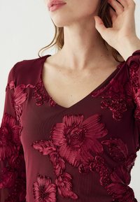 Burgundi värvi V-kaelusega topp, millel on keerukas lilleline tikand tekstuurilises kangas, rõhutades disaini kihiliste õite ja lehtedega.