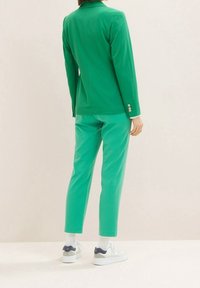 Personne debout, vue de dos, portant un blazer vert et un pantalon assorti avec des baskets blanches sur un fond clair.