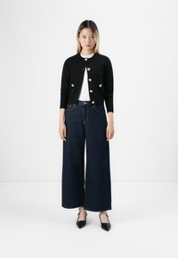 Femme debout portant un cardigan noir avec des boutons en forme de fleur, un haut blanc, un jean large bleu foncé et des chaussures noires à bout pointu sur fond blanc.