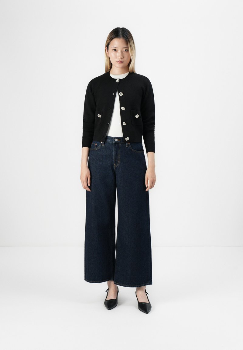 Femme debout portant un cardigan noir avec des boutons en forme de fleur, un haut blanc, un jean large bleu foncé et des chaussures noires à bout pointu sur fond blanc.