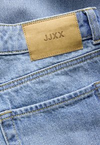 Jean en denim bleu clair avec un patch en cuir marron cousu au-dessus de la poche arrière embossé du nom de la marque JJXX.