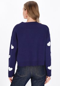 Temno modri skrajšan pulover z belimi srčastimi vzorci na rokavih, pletena tekstura in rebrast rob. Združitev s temno modrimi jeans hlačami.