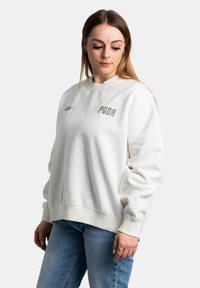 Felpa bianca con colletto a girocollo, polsini e orlo a costine, con testo e logo marrone chiaro "PGDR" sul davanti. Vestibilità casual con texture morbida.