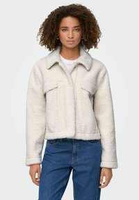 Donna che indossa una giacca di pile bianca con colletto alto e tasche frontali, abbinata a jeans blu, in piedi contro uno sfondo chiaro e neutro.