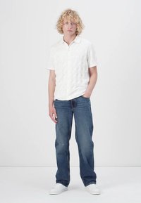 Witte shirt met korte mouwen en een gestructureerd patroon, gecombineerd met blauwe rechte jeans en witte sneakers. Model staat tegen een effene achtergrond.