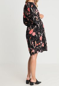 Schwarzes Blumenkleid mit langen Ärmeln, verziert mit rosa und orangefarbenen Blumen, aus fließendem Stoff, knielanger Schnitt, kombiniert mit schwarzen Blockabsatzschuhen.